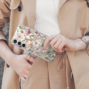 May include: A clear phone case featuring a floral design with white, pink, and yellow flowers. The case has a transparent background and a protective design. The phone case is held in front of a tan trench coat.