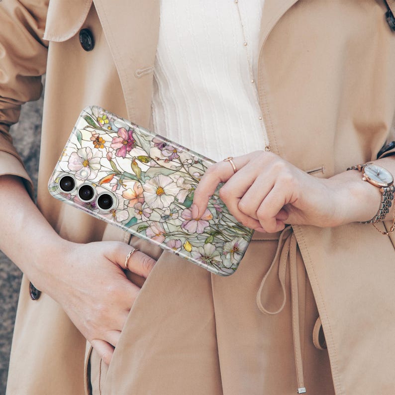 May include: A clear phone case featuring a floral design with white, pink, and yellow flowers. The case has a transparent background and a protective design. The phone case is held in front of a tan trench coat.
