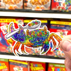 May include: A colorful, glittery crab holding a spoon full of rainbow cereal. The crab is sitting on a white background with a blue and white pattern. The text "Ryan McArthur" is written in black on the crab's shell.