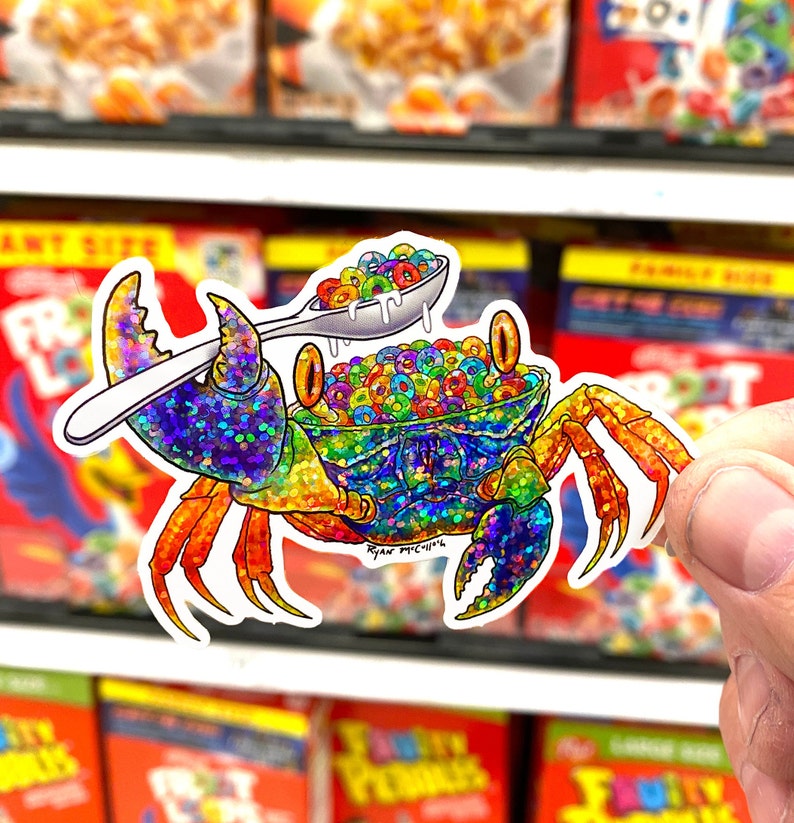 May include: A colorful, glittery crab holding a spoon full of rainbow cereal. The crab is sitting on a white background with a blue and white pattern. The text "Ryan McArthur" is written in black on the crab's shell.