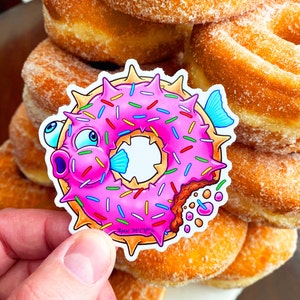 May include: A pink and white donut shaped like a pufferfish with sprinkles and a bite taken out of it. The donut is on top of a stack of donuts.