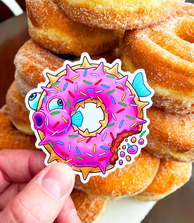 May include: A pink and white donut shaped like a pufferfish with sprinkles and a bite taken out of it. The donut is on top of a stack of donuts.