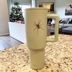 Puede incluir: Una taza de viaje verde claro con un gr&aacute;fico de ara&ntilde;a negra y un asa negra. La taza tiene una pajita negra y la palabra "slm" impresa en la parte inferior.