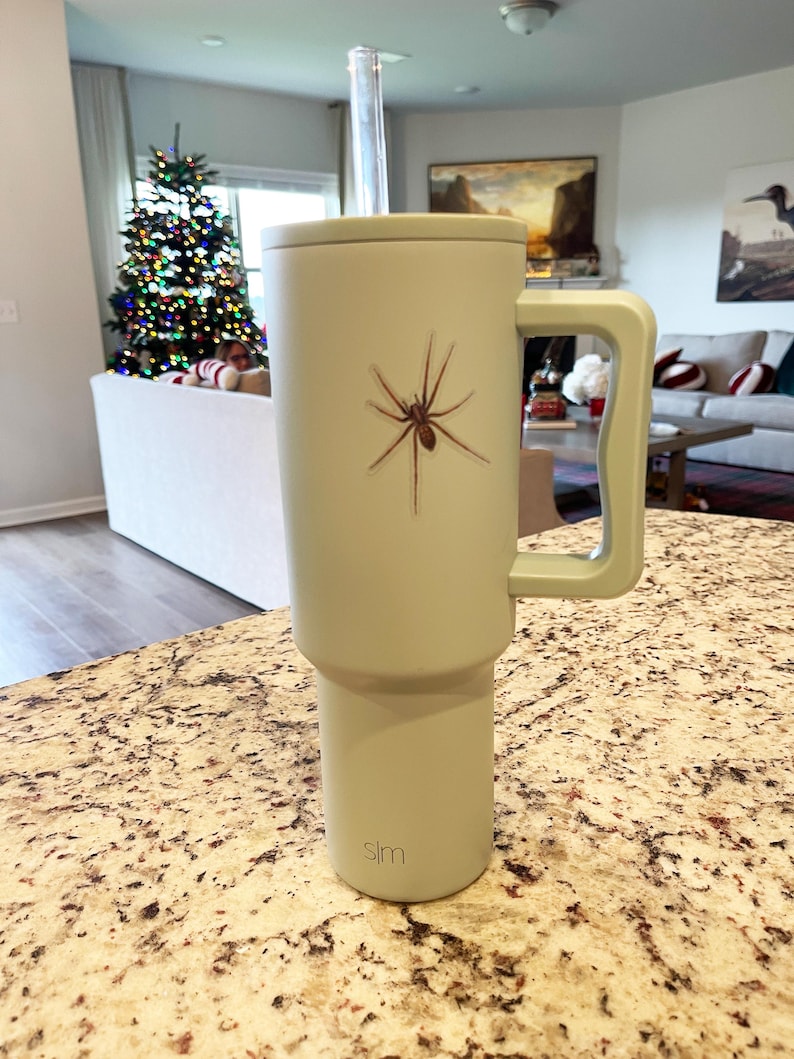 Puede incluir: Una taza de viaje verde claro con un gr&aacute;fico de ara&ntilde;a negra y un asa negra. La taza tiene una pajita negra y la palabra "slm" impresa en la parte inferior.