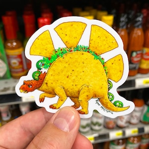 May include: A sticker of a cartoon dinosaur made of a taco shell. The dinosaur has a green jalapeno tail and a red and yellow head. The taco shell is filled with lettuce and cheese. The sticker is signed "Ryan McCulloch".