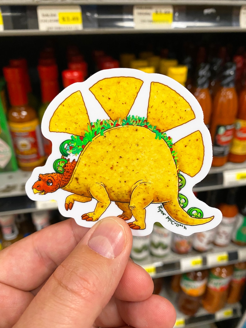 May include: A sticker of a cartoon dinosaur made of a taco shell. The dinosaur has a green jalapeno tail and a red and yellow head. The taco shell is filled with lettuce and cheese. The sticker is signed "Ryan McCulloch".