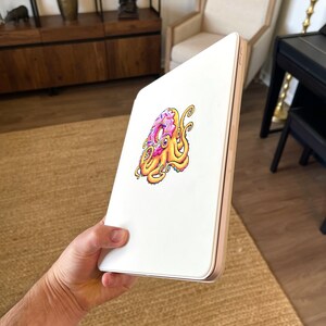 May include: A white iPad case with a colorful illustration of an octopus holding a donut. The donut is pink with white sprinkles and the octopus is yellow with purple tentacles.