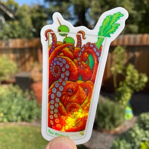 May include: A sticker featuring a cocktail glass filled with an octopus. The octopus is orange and red with blue accents. The drink is garnished with green olives and a celery stalk. The artist's signature is at the bottom.
