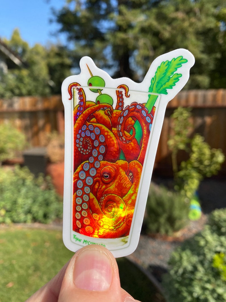 May include: A sticker featuring a cocktail glass filled with an octopus. The octopus is orange and red with blue accents. The drink is garnished with green olives and a celery stalk. The artist's signature is at the bottom.