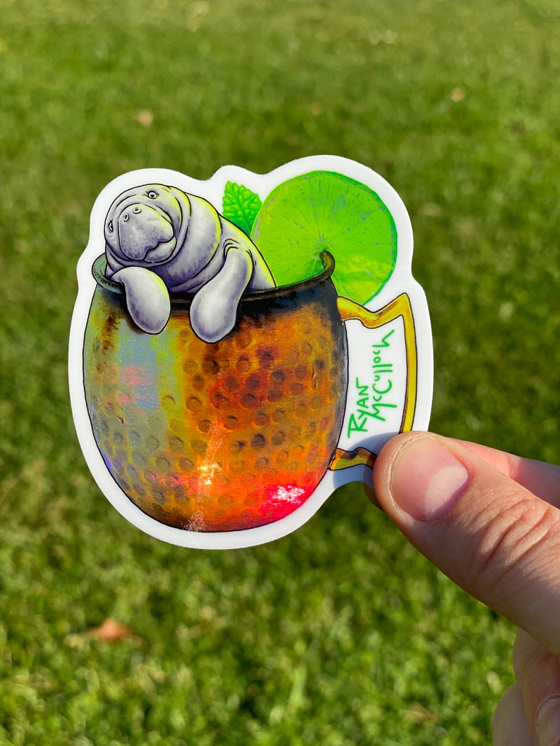 May include: A sticker featuring a manatee relaxing in a copper mug. The mug is adorned with a lime slice and mint sprig. The sticker has the text "Ryan McCulloch" on the handle.