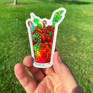 May include: A sticker featuring an octopus inside a cocktail glass, with a green olive and celery garnish. The octopus is orange and red, with iridescent highlights. The sticker has a white border and the artist's signature.