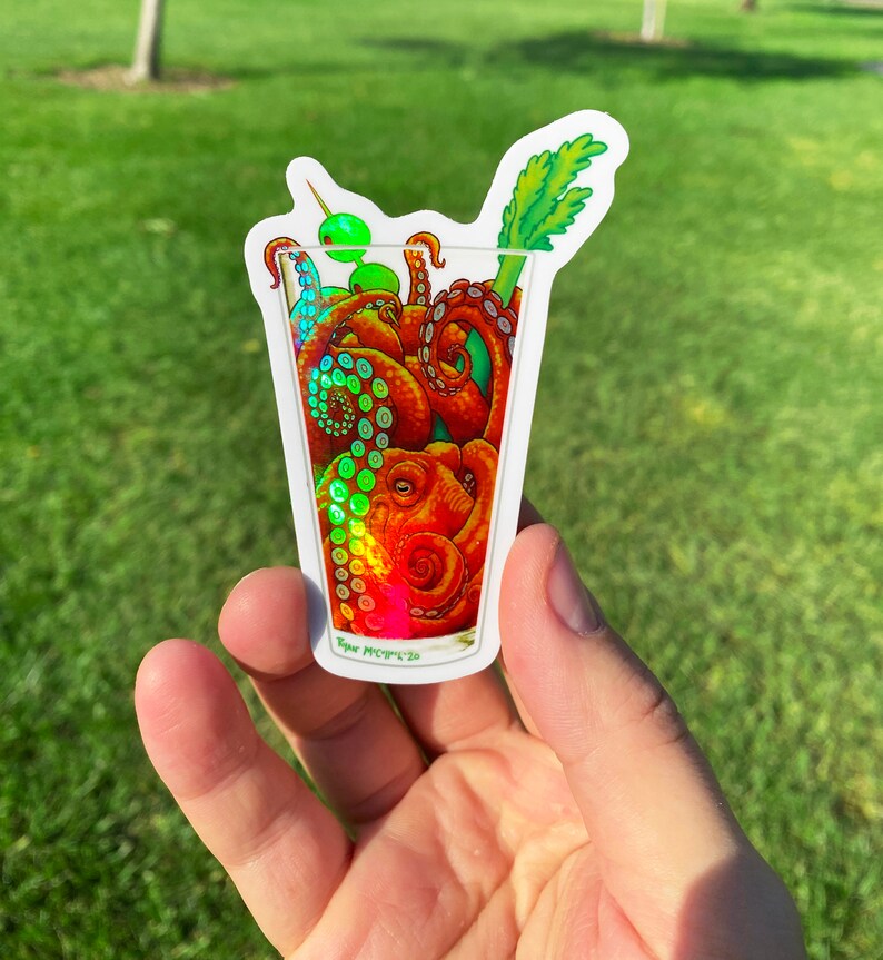 May include: A sticker featuring an octopus inside a cocktail glass, with a green olive and celery garnish. The octopus is orange and red, with iridescent highlights. The sticker has a white border and the artist's signature.