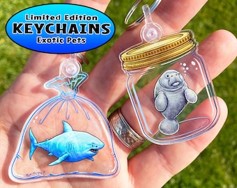 May include: Two acrylic keychain charms featuring exotic animals. The first charm is a clear plastic bag with a blue and white shark inside. The second charm is a clear glass jar with a manatee inside. Both charms have a gold top and a silver ring.