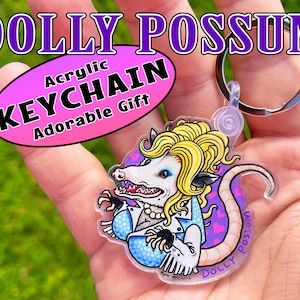 May include: Acrylic keychain featuring a cartoon possum with blonde hair, wearing a blue and white polka dot top, a pearl necklace, and a pink heart on her tail. The keychain is labeled "DOLLY POSSUM" and "Acrylic Keychain Adorable Gift".
