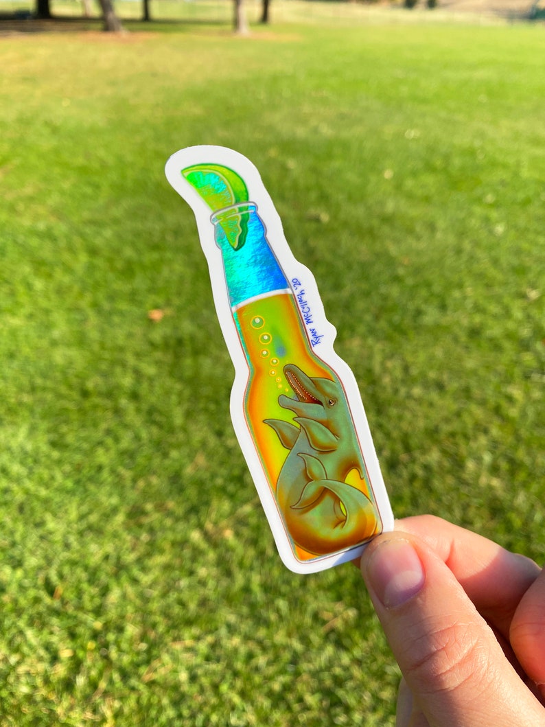 May include: A sticker in the shape of a beer bottle with a lime wedge on top. Inside the bottle are two dolphins. The bottle has a gradient of colors, including blue, yellow, and green. The text "Ryan Mcclintock 20" is on the bottle.