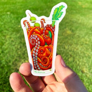 May include: A sticker featuring a cocktail glass filled with orange octopus tentacles, green olives, and a celery stalk. The sticker has a white border and the artist's signature.