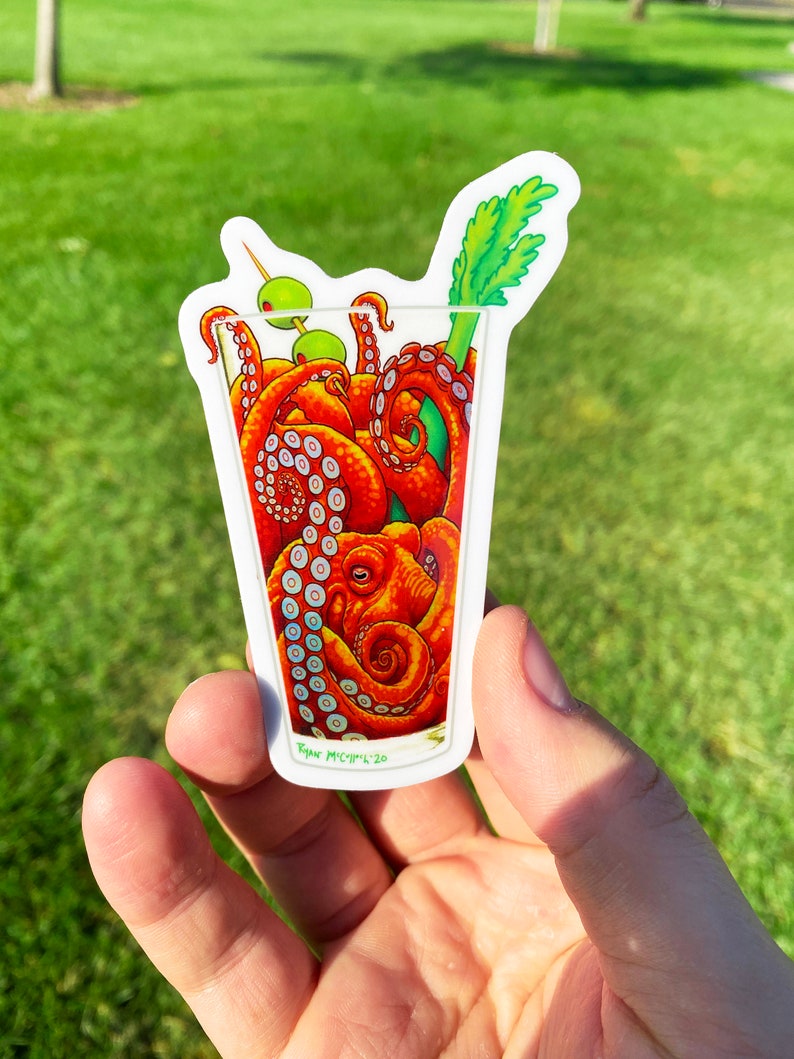 May include: A sticker featuring a cocktail glass filled with orange octopus tentacles, green olives, and a celery stalk. The sticker has a white border and the artist's signature.