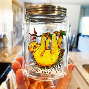 May include: A glass jar with a silver lid features a cartoon illustration of a sloth hanging upside down from a branch. The sloth is yellow with a brown face and is surrounded by colorful vegetables and red peppers.