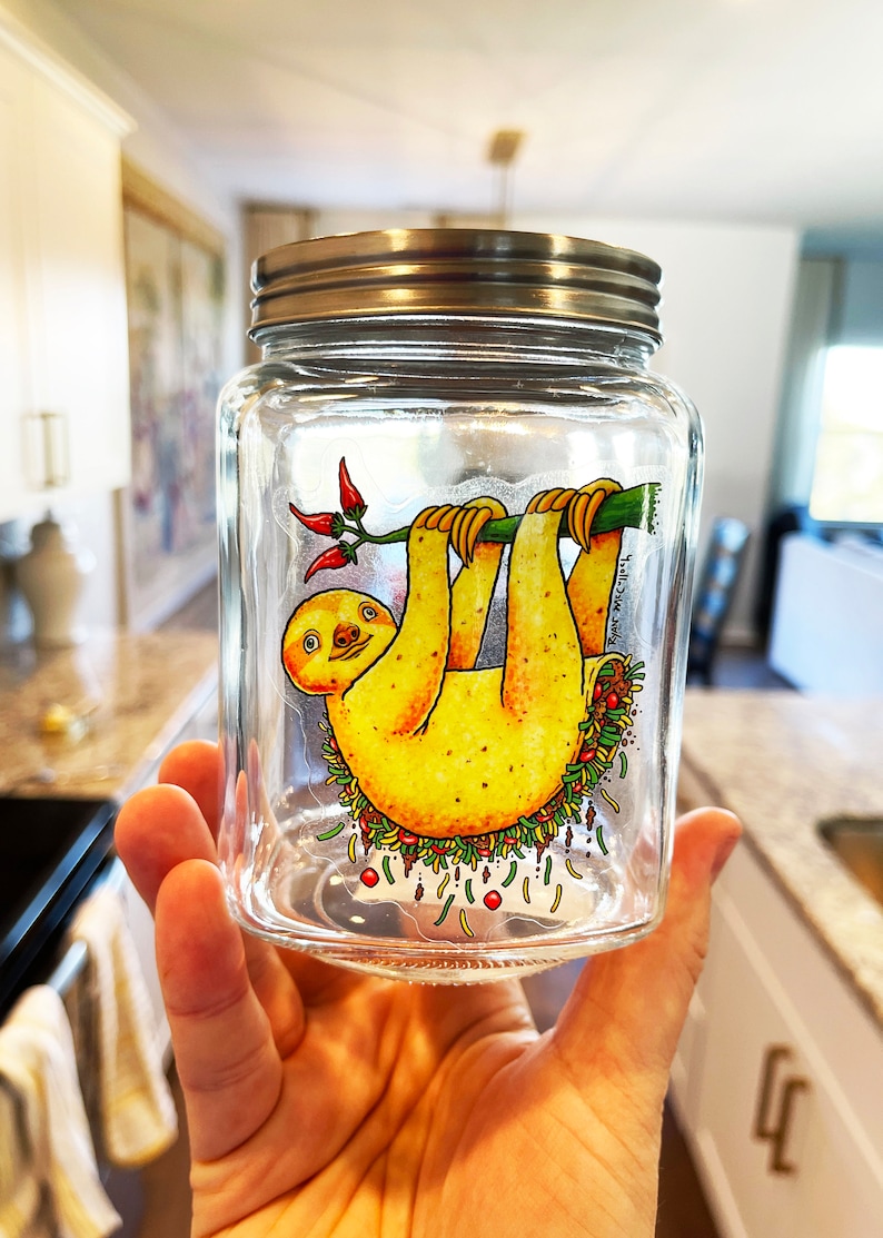 May include: A glass jar with a silver lid features a cartoon illustration of a sloth hanging upside down from a branch. The sloth is yellow with a brown face and is surrounded by colorful vegetables and red peppers.