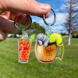 May include: Two keychains featuring a glass of bloody mary with an octopus and a manatee in a copper mug with a lime wedge. The manatee keychain has the text "Rum Punch" on the side.