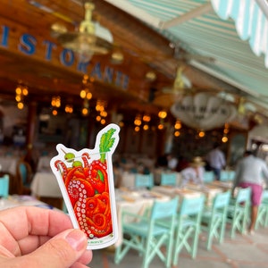 May include: A sticker featuring a cartoon illustration of a cocktail glass filled with an octopus. The octopus is red with curled tentacles, olives, and a celery stalk garnish. The background is a blurred outdoor restaurant scene.