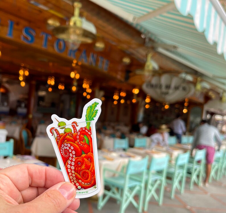 May include: A sticker featuring a cartoon illustration of a cocktail glass filled with an octopus. The octopus is red with curled tentacles, olives, and a celery stalk garnish. The background is a blurred outdoor restaurant scene.