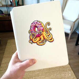 May include: A white notebook with a sticker of a pink and yellow donut with sprinkles being held by an octopus with yellow tentacles.