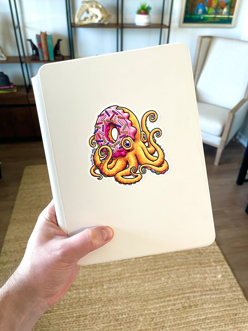 May include: A white notebook with a sticker of a pink and yellow donut with sprinkles being held by an octopus with yellow tentacles.