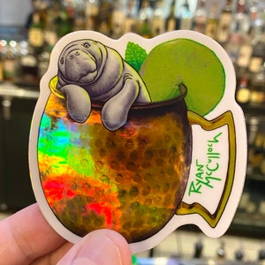 May include: A sticker featuring a cartoon manatee relaxing in a copper mug, with a lime wedge and mint sprig. The mug has a textured, holographic finish. The sticker is labeled "Ryan McCulloch" in green text.