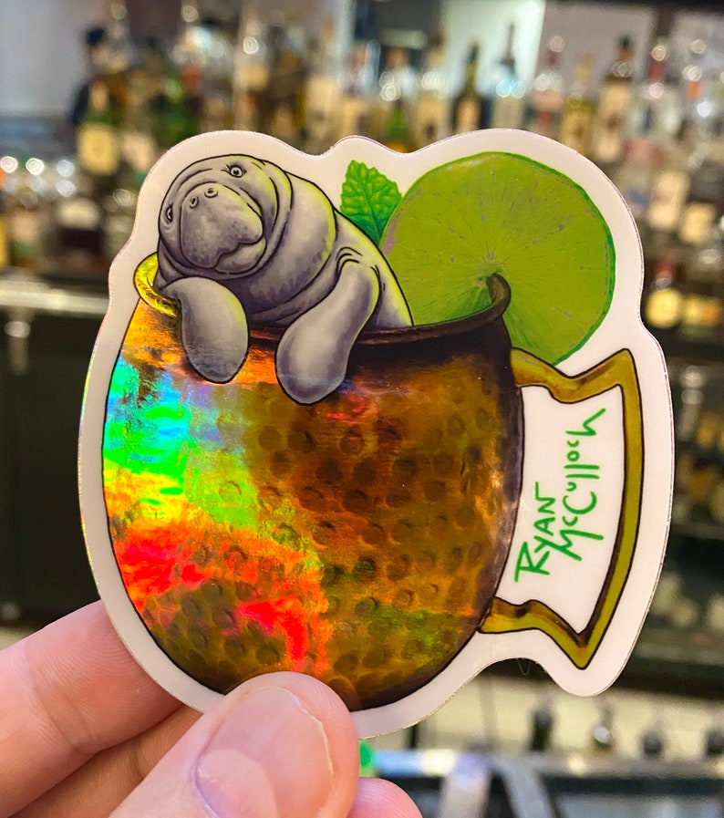 May include: A sticker featuring a cartoon manatee relaxing in a copper mug, with a lime wedge and mint sprig. The mug has a textured, holographic finish. The sticker is labeled "Ryan McCulloch" in green text.