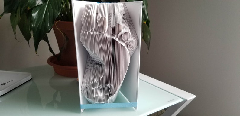May include: A white paper book folded into the shape of a footprint. The book is standing upright on a white table with a blue ribbon around the bottom.