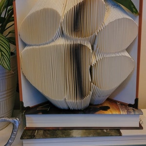 May include: A decorative book sculpture with pages folded into a flower shape. The book is propped up on a stack of other books. The sculpture is beige and brown, with a plant in the background.