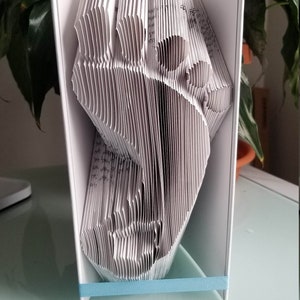 May include: A white book folded into a three-dimensional shape resembling a bird. The book is standing on a white table with a light blue ribbon around the base.