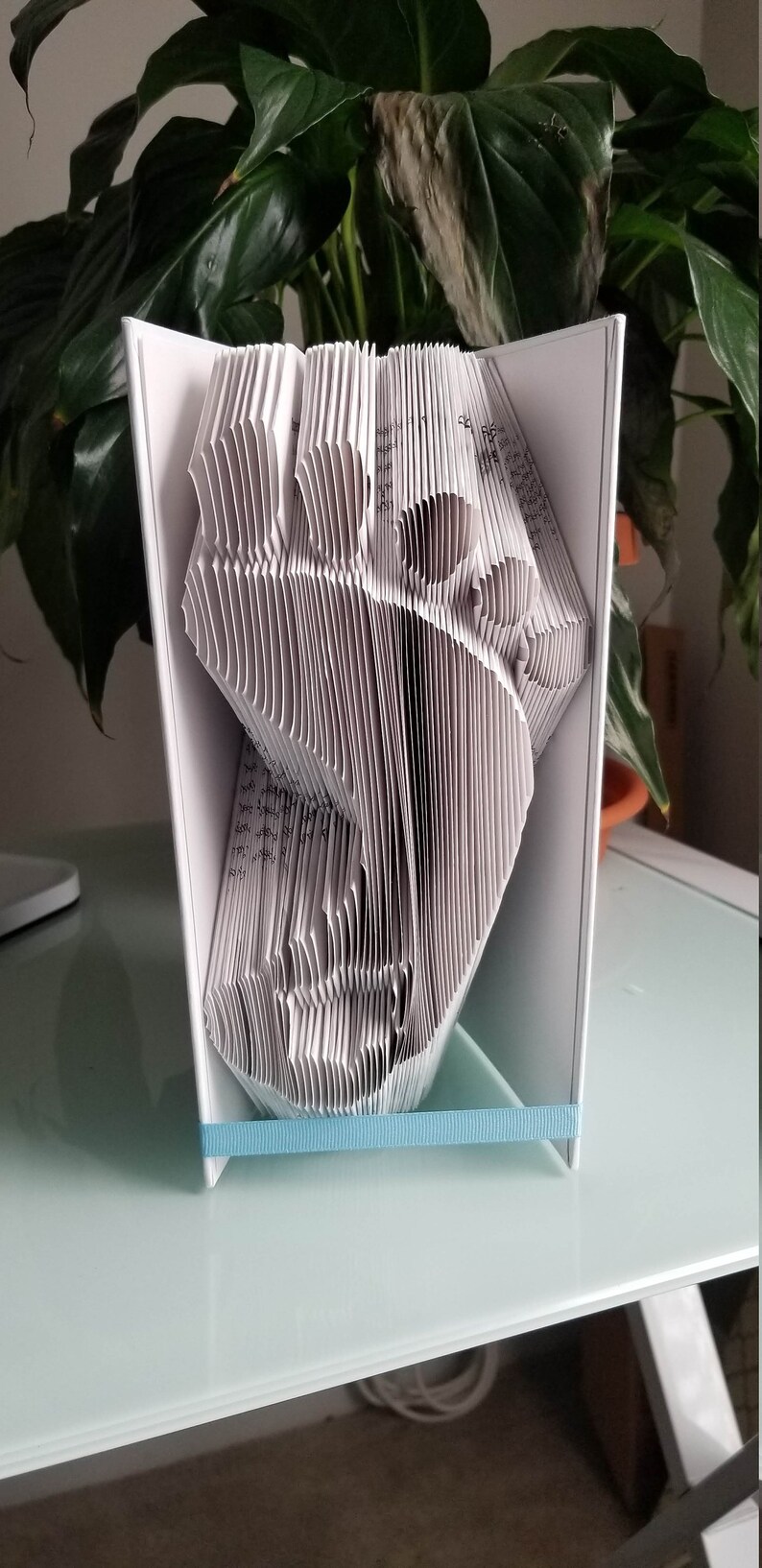 May include: A white book folded into a three-dimensional shape resembling a bird. The book is standing on a white table with a light blue ribbon around the base.