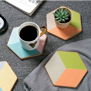 May include: A white coffee cup with a gold handle and a heart design sits on a geometric coaster with blue, pink, and tan sections. Other coasters with green, orange, and tan sections are also visible. A small potted succulent is on one coaster.
