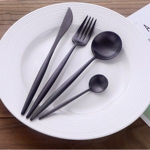 May include: A set of matte black silverware, including a knife, fork, spoon, and teaspoon, arranged on a white plate with a ridged design. The utensils have long, slender handles and are made of a dark metal.