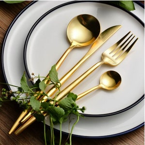 May include: A set of gold-colored flatware, including a spoon, knife, fork, and small spoon, arranged on a white plate with a blue rim. Green sprigs of leaves are placed near the silverware. The flatware has a shiny, reflective surface.