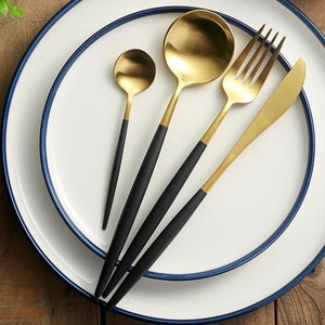 May include: A set of gold-colored flatware with black handles, including a spoon, teaspoon, fork, and knife, arranged on a white plate with a blue rim. The flatware has a modern design.