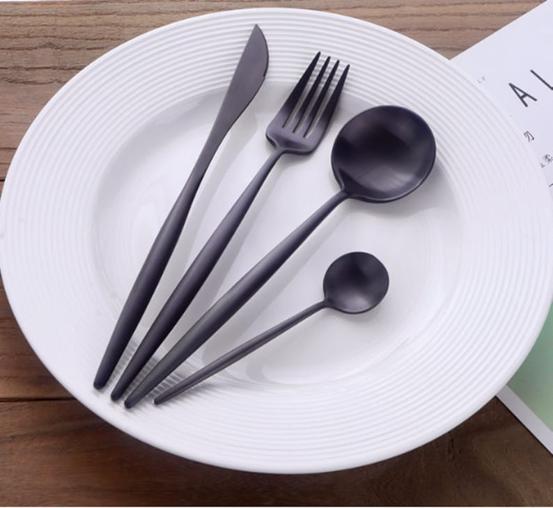 May include: A set of matte black silverware, including a knife, fork, spoon, and teaspoon, arranged on a white plate with a ridged design. The utensils have long, slender handles and are made of a dark metal.