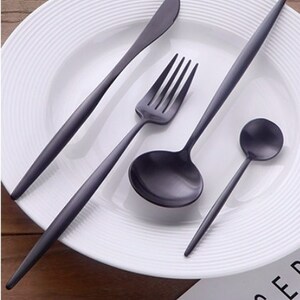 May include: A matte black flatware set, including a knife, fork, spoon, and ladle, is arranged on a white plate. The utensils have a sleek, modern design with long, slender handles. The plate has a ridged rim, and the background is a wooden surface.
