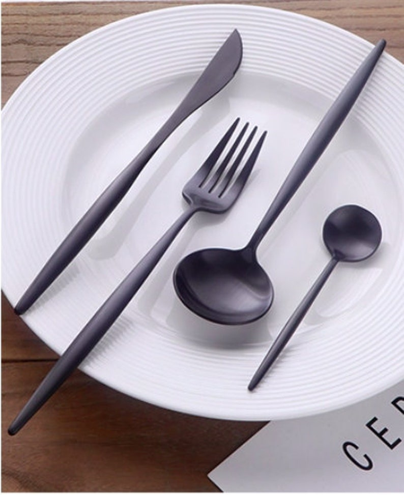 May include: A matte black flatware set, including a knife, fork, spoon, and ladle, is arranged on a white plate. The utensils have a sleek, modern design with long, slender handles. The plate has a ridged rim, and the background is a wooden surface.