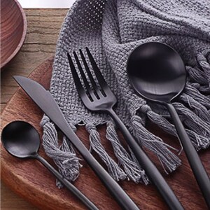 May include: A set of black matte finish silverware including a knife, fork, spoon, and teaspoon, arranged on a wooden cutting board. A gray fringed cloth is in the background. The silverware has a sleek, modern design.