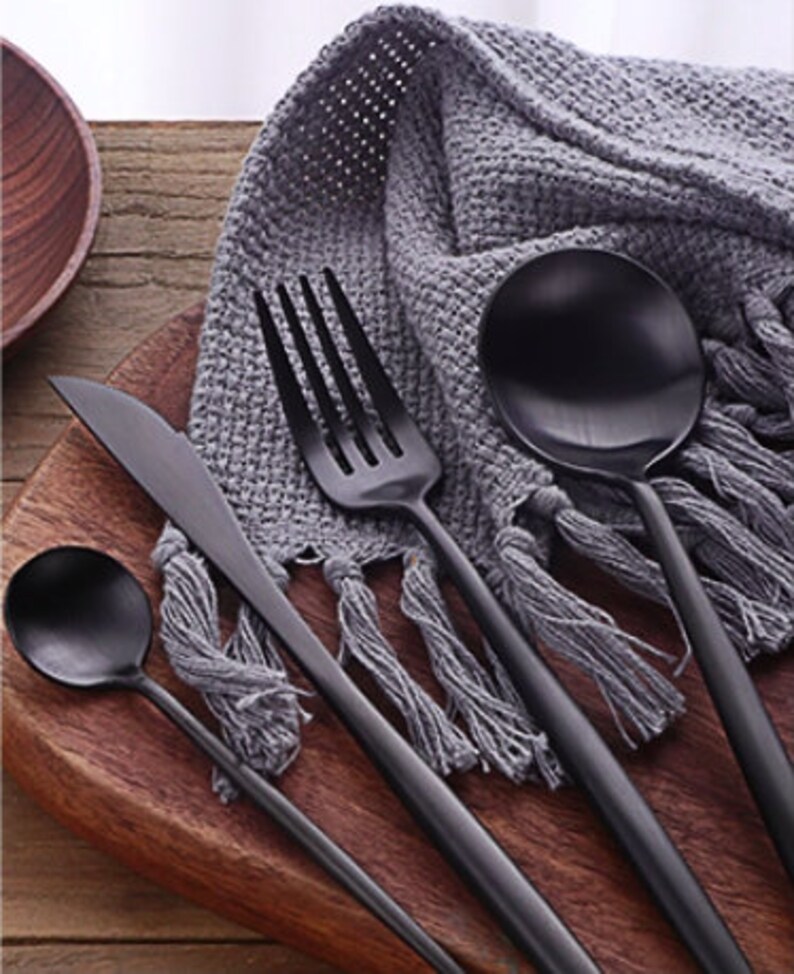 May include: A set of black matte finish silverware including a knife, fork, spoon, and teaspoon, arranged on a wooden cutting board. A gray fringed cloth is in the background. The silverware has a sleek, modern design.