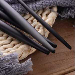 May include: Close-up of black matte finish utensils, possibly chopsticks or serving pieces. The utensils are arranged on a woven straw mat with a gray fabric in the background. The dark color of the utensils contrasts with the lighter background.