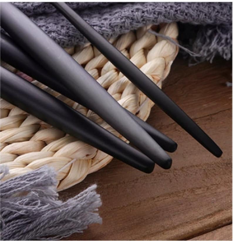 May include: Close-up of black matte finish utensils, possibly chopsticks or serving pieces. The utensils are arranged on a woven straw mat with a gray fabric in the background. The dark color of the utensils contrasts with the lighter background.