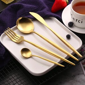 May include: A set of gold-colored flatware, including a fork, spoon, knife, and teaspoon, arranged on a white rectangular plate. The flatware has a sleek, modern design. A cup of tea and a strawberry are in the background.