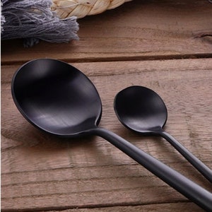 May include: Two black serving spoons on a wooden surface. The spoons have a matte finish and a simple, modern design. The larger spoon is oval-shaped, and the smaller spoon is round.