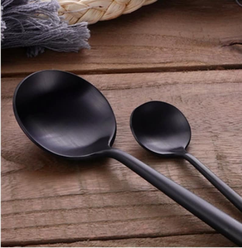 May include: Two black serving spoons on a wooden surface. The spoons have a matte finish and a simple, modern design. The larger spoon is oval-shaped, and the smaller spoon is round.