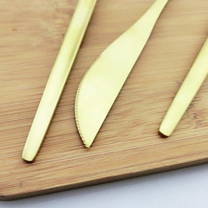 May include: Close-up of three pieces of gold-colored flatware on a wooden surface. The set includes a knife with a serrated edge and two utensils with elongated, tapered handles. The flatware has a matte finish.