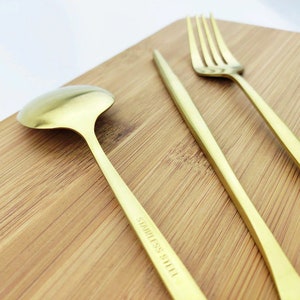 May include: A close-up of gold-colored stainless steel cutlery set on a wooden cutting board. The set includes a spoon, a knife, and a fork. The spoon has a rounded bowl, and the knife has a pointed tip. The fork has four tines. The words "STAINLESS STEEL" are printed on the knife.
